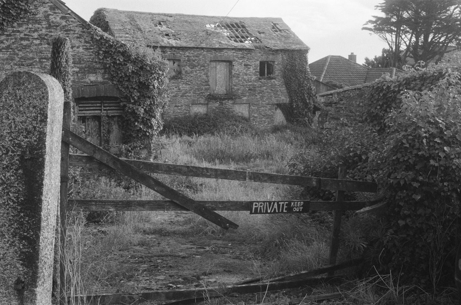 Black and white photograph of an old stone building with ivy, a broken wooden roof section, a stone gatepost, and a wooden gate with a sign reading PRIVATE. Trees and shrubs surround the property.