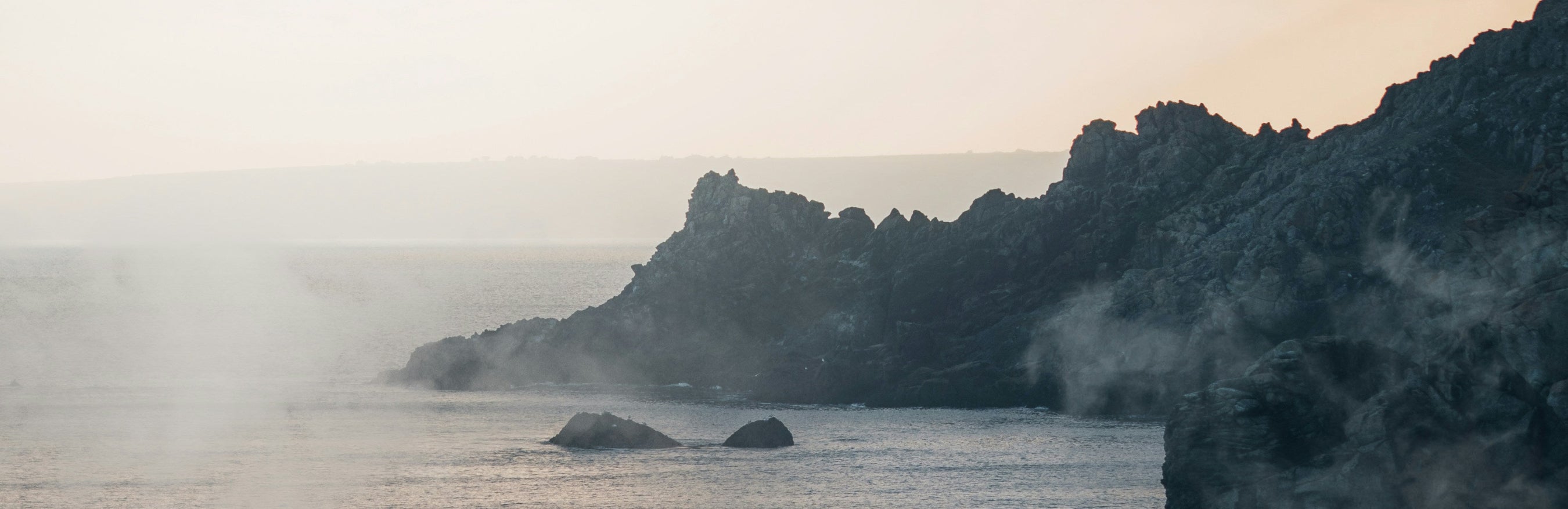 A misty coastal seascape with dark rocky cliffs along the right side, calm water in the foreground, and a bright hazy sky with low clouds.