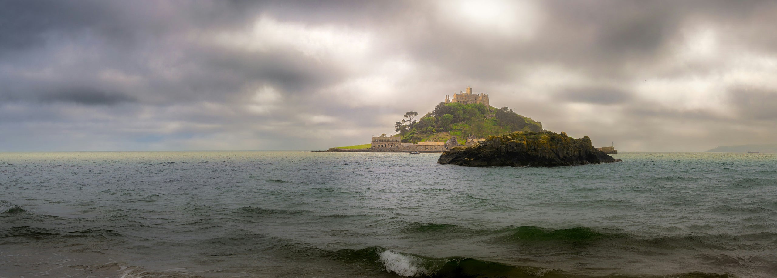A scenic coastal landscape showing a small island with a castle-like structure under a dramatic cloudy sky, with choppy water in the foreground.