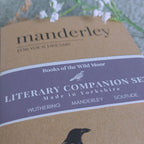 A close-up of a brown paper book cover for the Manderley Literary Companion Set, featuring a dark purple label with white text and a small bird illustration. The cover includes the title 'manderley' and references to Wuthering, Manderley, and Solitude.