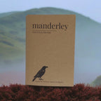 A square kraft-paper style book or notebook cover standing upright, printed with the word "manderley" and smaller text "FOR YOUR DREAMS," plus a black bird silhouette near the bottom. The product is photographed outdoors against a blurred green hill and pale sky, with reddish foreground foliage.