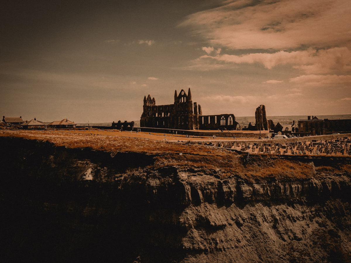 A sepia-toned landscape photograph of a large stone abbey ruin situated on a grassy cliffside under a cloudy sky.
