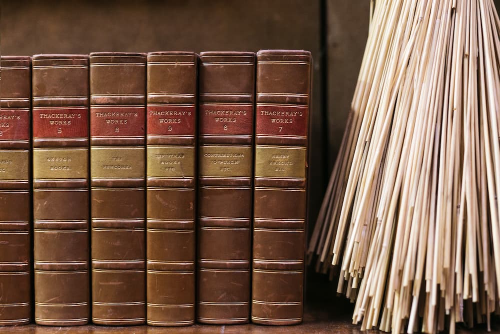A row of vintage leather-bound books with brown spines and red-and-gold title bands, standing beside a bundle of loose pages or sheets.